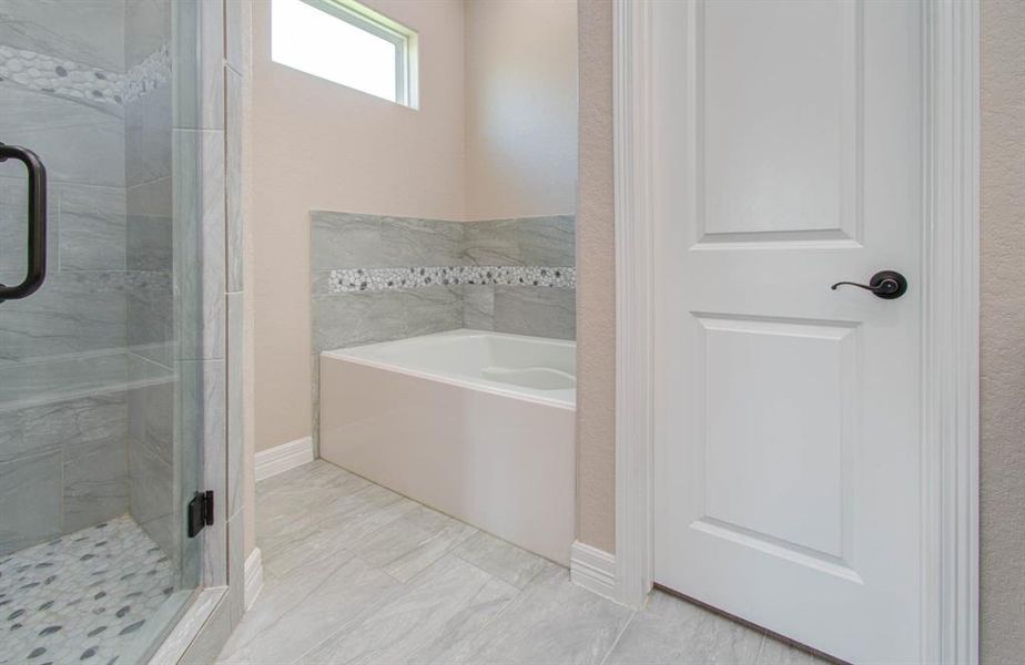 This bathroom features modern grey tile flooring, a spacious walk-in shower with a glass door, and an accent-tiled bathtub. The room is well-lit with a frosted window and includes a white door with dark hardware.