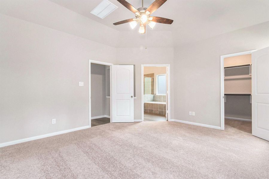 This spacious bedroom features soft carpet flooring, neutral walls, and a ceiling fan. It includes a walk-in closet and en suite bathroom with a large bathtub, adding convenience and luxury.