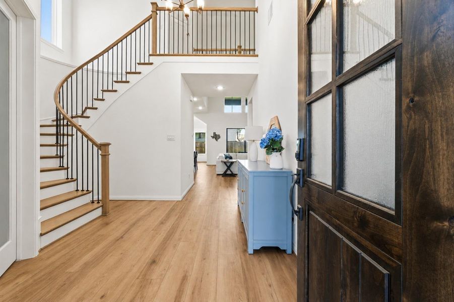 This inviting entryway features a wooden door, signature curved staircase with elegant railings, and bright, airy interiors with wood look vinyl flooring.