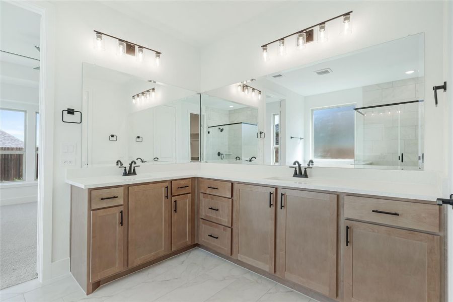 Master bathroom with double vanity, wood cabinets, white countertops, and large mirror with glass shower in background.
