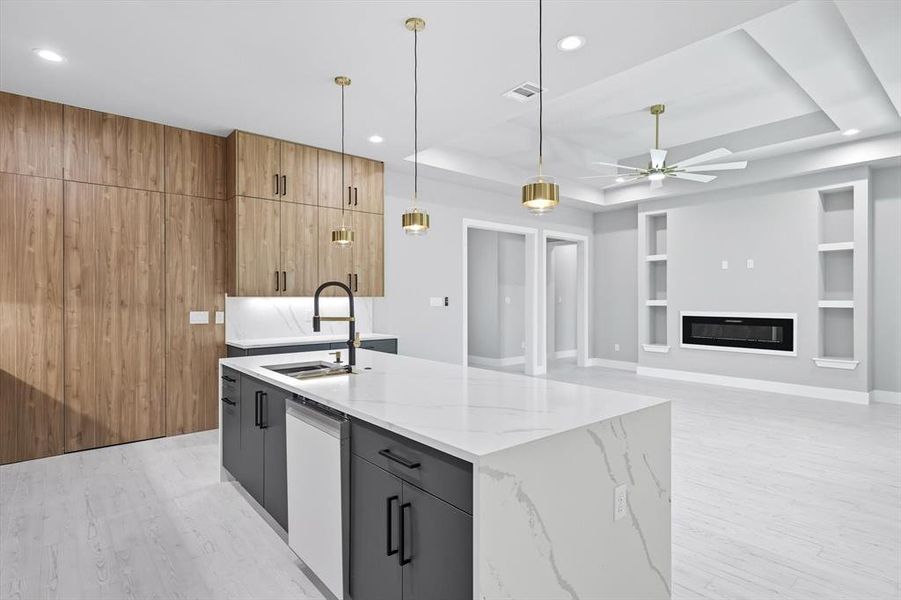 Kitchen with pendant lighting, an island with sink, built in shelves, light stone countertops, and a tray ceiling