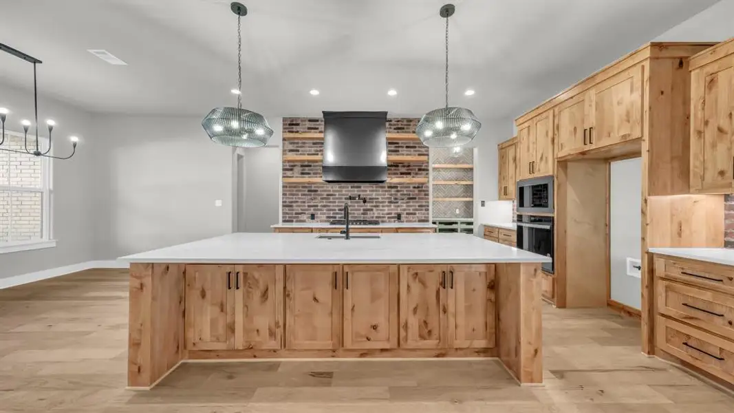Open-concept kitchen featuring a large central island with integrated sink, light wood cabinetry, white countertops, and a brick-patterned backsplash Open-concept kitchen featuring a large central island with integrated sink, light wood cabinetry, white countertops, and a brick-patterned backsplash