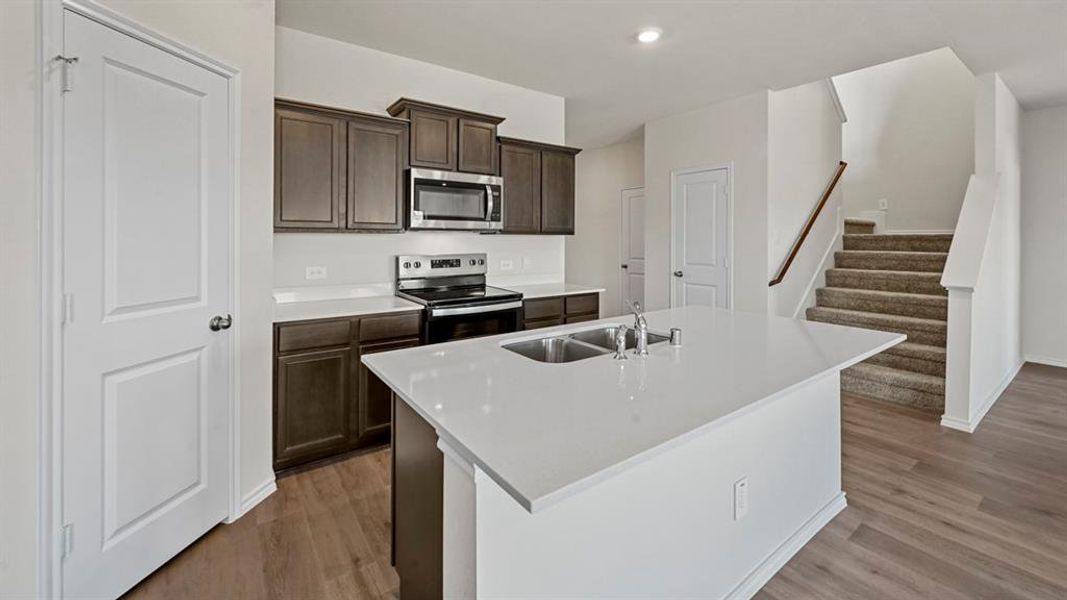 Kitchen featuring dark brown cabinets, appliances with stainless steel finishes, a kitchen island with sink, light wood-style floors, and light stone countertops