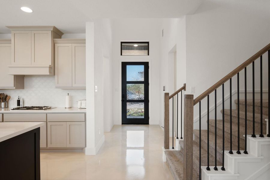 A striking modern entryway showcases a sleek black door with textured glass, flanked by a contemporary kitchen and a carpeted staircase with a chic railing. A striking modern entryway showcases a sleek black door with textured glass, flanked by a contemporary kitchen and a carpeted staircase with a chic railing.