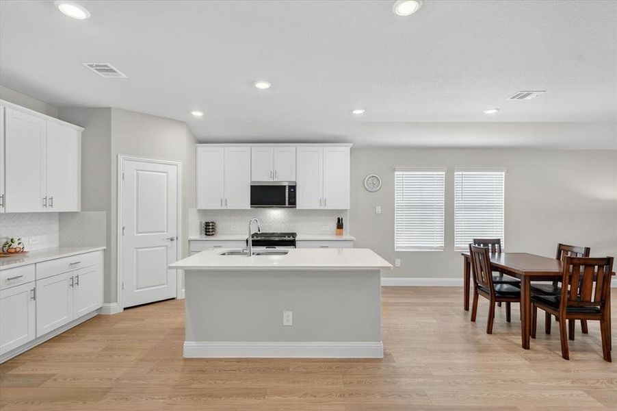 Kitchen featuring white cabinetry, light-toned countertops, a central island with a sink, stainless steel appliances, and wood-finish flooring Kitchen featuring white cabinetry, light-toned countertops, a central island with a sink, stainless steel appliances, and wood-finish flooring