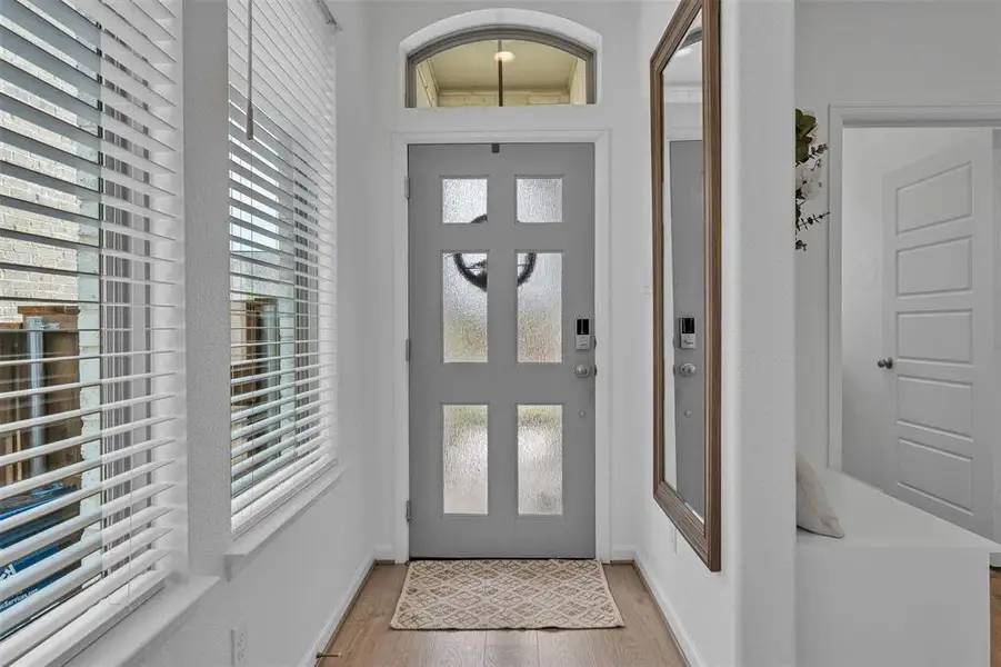 Entryway featuring a grey paneled door with frosted glass inserts, an arched transom window, wood-finish flooring, and white window blinds Entryway featuring a grey paneled door with frosted glass inserts, an arched transom window, wood-finish flooring, and white window blinds