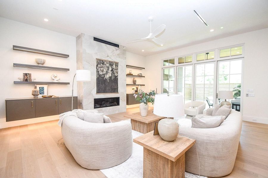 This bright living room features a modern, open design with light wood flooring, a sleek fireplace, and large windows for ample natural light. It includes comfortable seating, minimalist shelving, and a neutral color palette, creating a serene and inviting atmosphere.