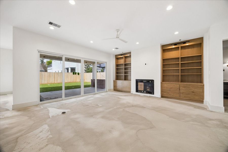 Spacious open concept living room designed for both comfort and entertaining, anchored by a sleek fireplace and flanked by custom built in shelving with warm wood finishes. Expansive sliding glass doors create a seamless connection to the covered patio and backyard, filling the space with natural light. Clean lines, recessed lighting, and a modern ceiling fan enhance the airy feel, while the thoughtful layout offers flexibility for a variety of furniture arrangements and everyday living.