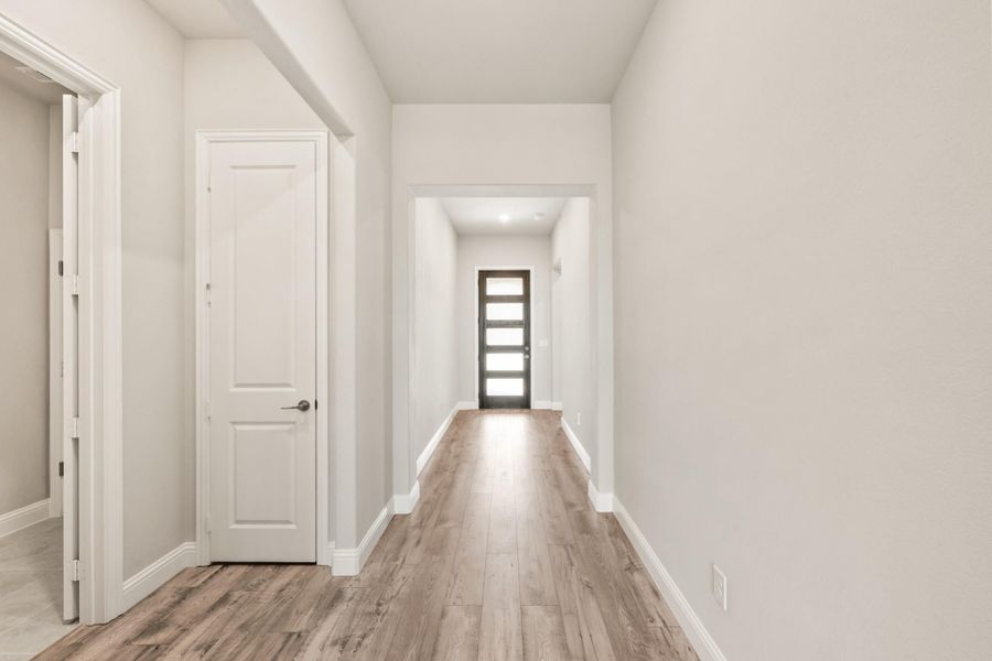 Hallway with light hardwood floors, white walls, and a dark glass front door at the end. Hallway with light hardwood floors, white walls, and a dark glass front door at the end.