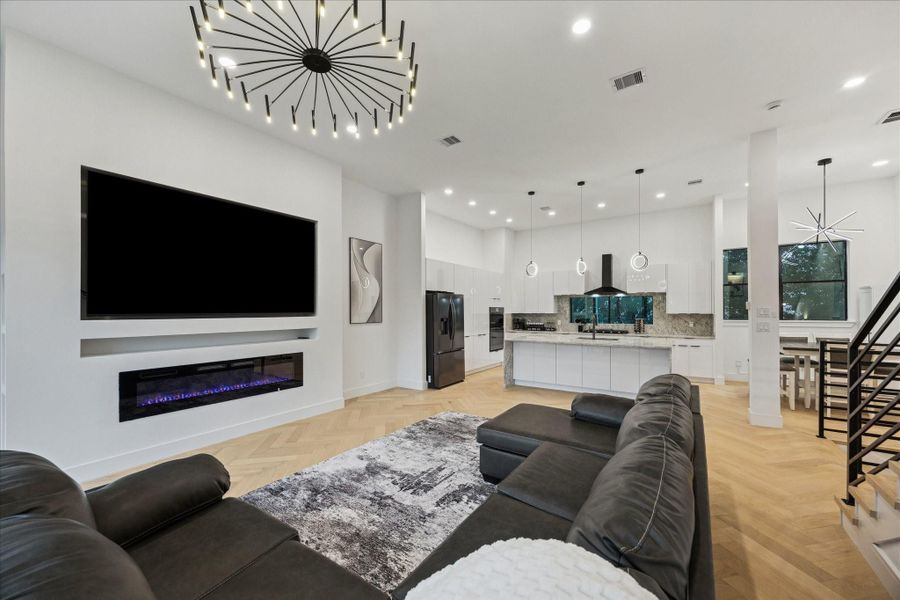 Open-concept living area featuring white oak herringbone flooring, dramatic designer lighting, and a sleek electric fireplace with a spacious media wall.