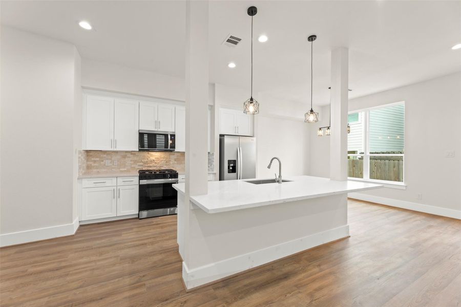 This modern kitchen features sleek white cabinetry, stainless steel appliances, and a spacious island with pendant lighting. The open layout and large window create a bright, inviting space with warm flooring.