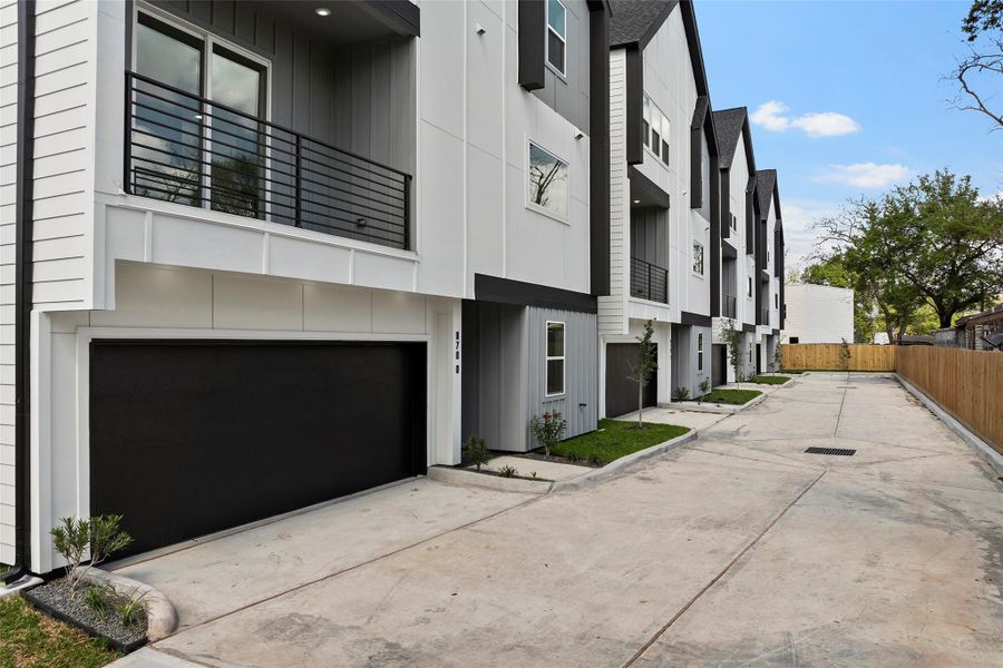 Modern townhomes with sleek black and white exteriors, featuring spacious garages and balconies. The driveway is wide and well-paved, lined with young trees and a privacy fence.