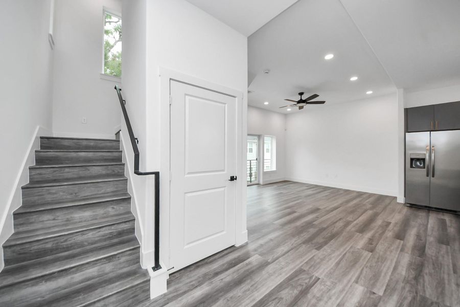 Modern open-concept living space featuring sleek wood flooring, recessed lighting, and a ceiling fan. The kitchen area includes stainless steel appliances, and there's a staircase leading to the upper level. Natural light streams through the windows, enhancing the bright, airy feel. Modern open-concept living space featuring sleek wood flooring, recessed lighting, and a ceiling fan. The kitchen area includes stainless steel appliances, and there's a staircase leading to the upper level. Natural light streams through the windows, enhancing the bright, airy feel.