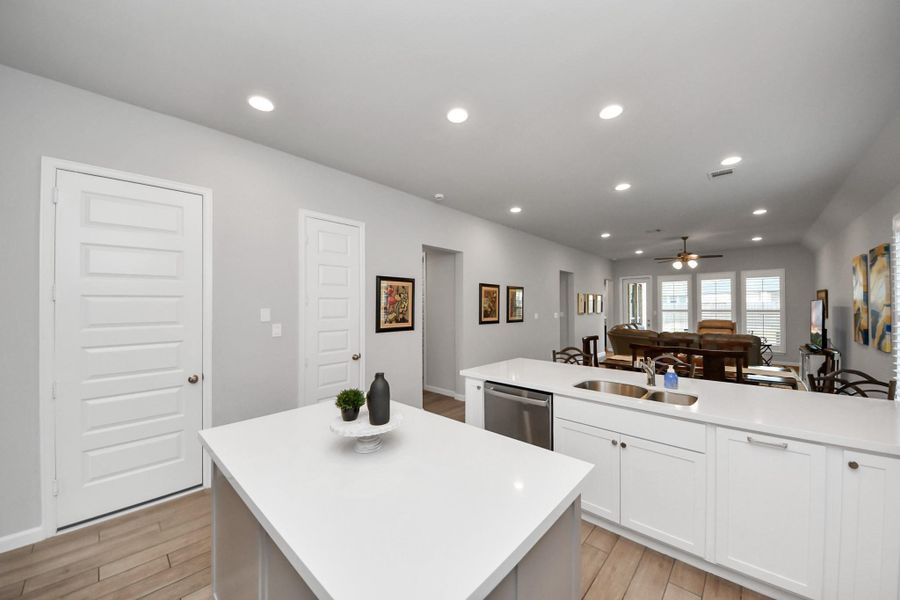This modern open-concept kitchen and living area features sleek white cabinetry, a central island, and recessed lighting. The space flows into a cozy living room with large windows, allowing for plenty of natural light.