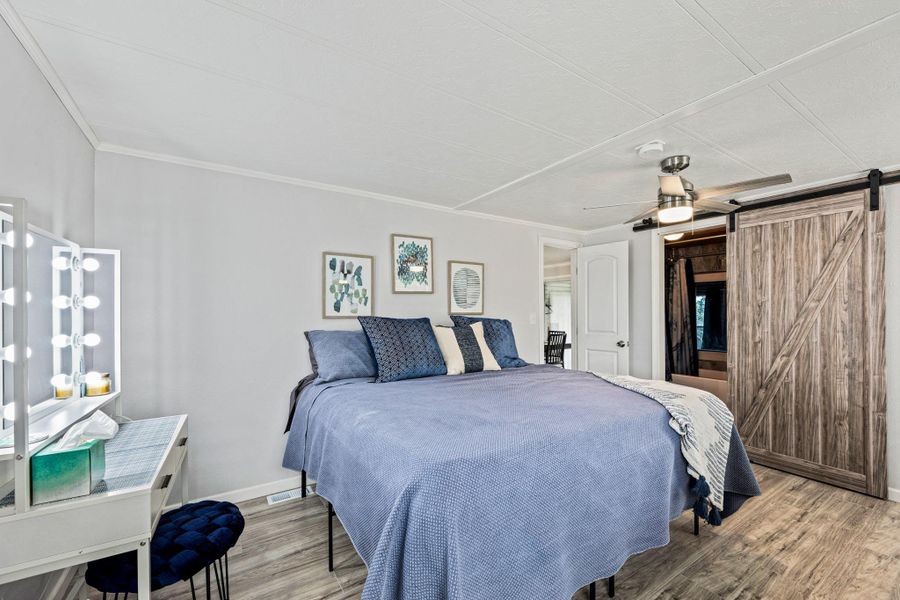 Spacious bedroom featuring wood-finish flooring, a ceiling fan with integrated lighting, a stylish barn door, and bright, neutral wall tones
