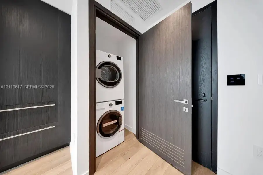 Laundry area featuring a stackable washer and dryer, wood-finish flooring, custom dark-toned cabinetry, and minimalist doors with linear detailing Laundry area featuring a stackable washer and dryer, wood-finish flooring, custom dark-toned cabinetry, and minimalist doors with linear detailing