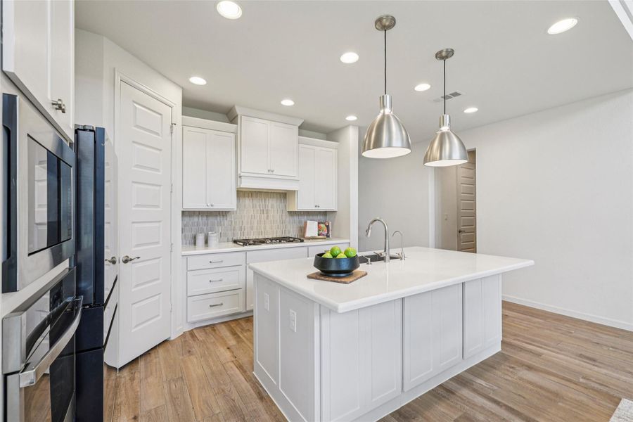 The kitchen features a central island with a sink, white cabinetry, a tiled backsplash, built-in ovens, and recessed lighting