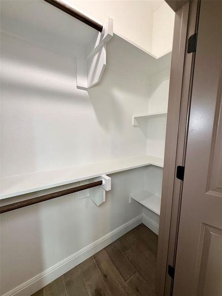 Walk-in closet featuring wood-finish flooring, white baseboards, white walls, built-in white shelving, and a dark-finish hanging rod, closets for bedroom 2 and 3 have same closet layout and custom shelves Walk-in closet featuring wood-finish flooring, white baseboards, white walls, built-in white shelving, and a dark-finish hanging rod, closets for bedroom 2 and 3 have same closet layout and custom shelves