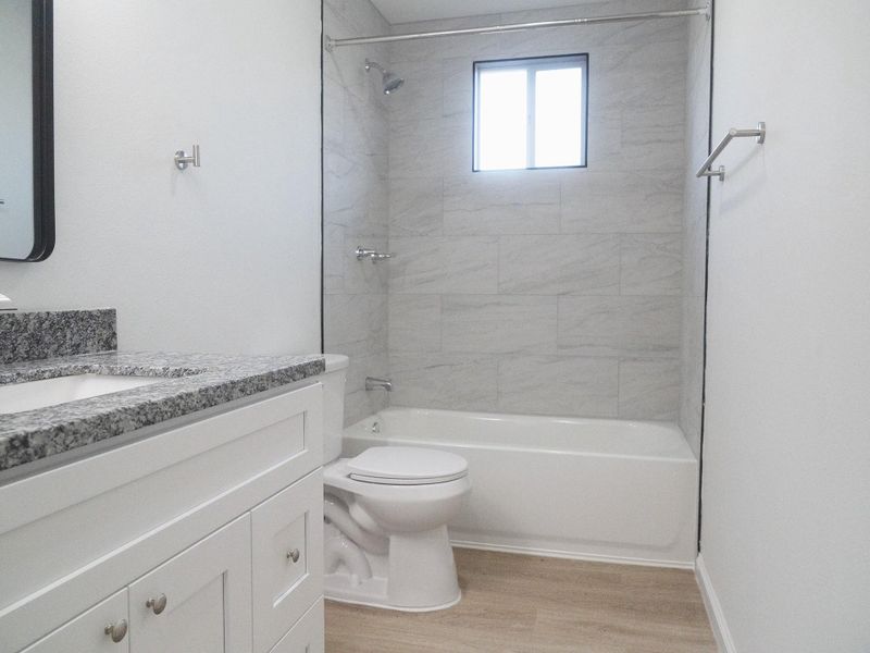 This bathroom features a modern design with a granite countertop, white cabinetry, and a tiled bath/shower area. It has a window for natural light and a clean, neutral color palette.