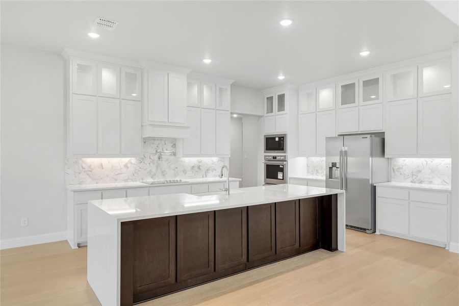 Large kitchen with white cabinets, marble backsplash, dark wood island, and stainless steel appliances on light hardwood floors