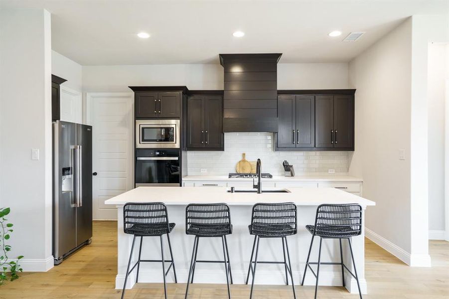 Kitchen with stainless steel appliances, light wood-style flooring, light countertops, a kitchen island with sink, and backsplash Kitchen with stainless steel appliances, light wood-style flooring, light countertops, a kitchen island with sink, and backsplash
