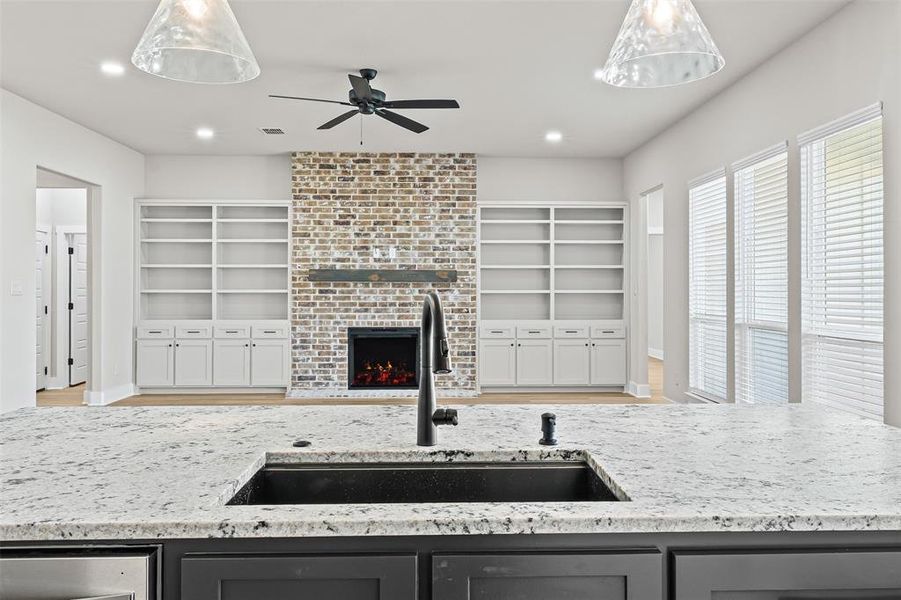 Kitchen featuring light stone countertops, a brick fireplace, a ceiling fan, and built in shelves Kitchen featuring light stone countertops, a brick fireplace, a ceiling fan, and built in shelves