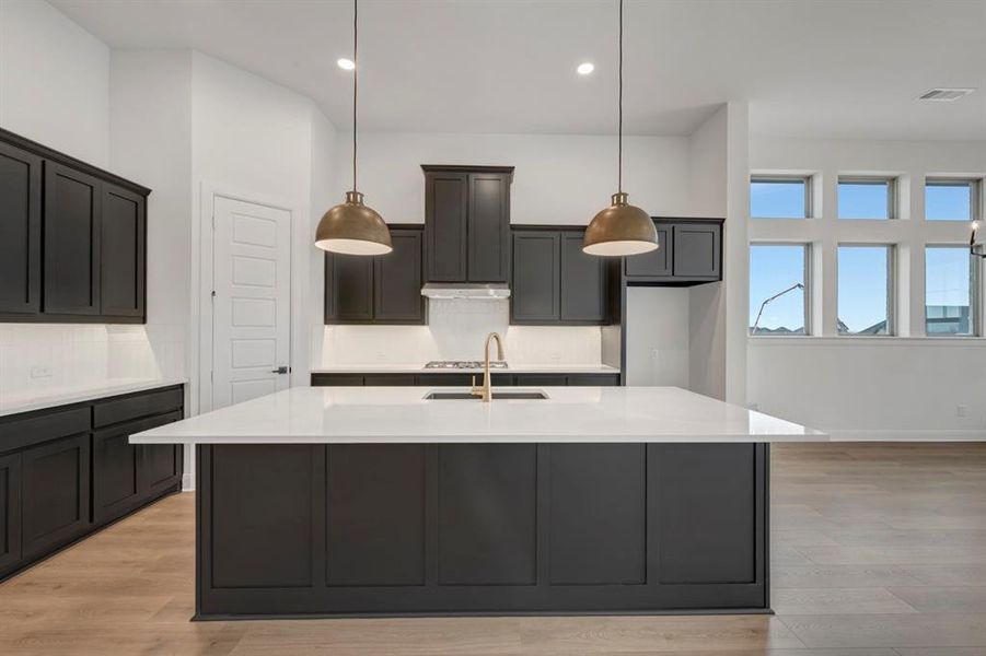 Kitchen with light stone counters, an island with sink, light wood-type flooring, and decorative light fixtures
