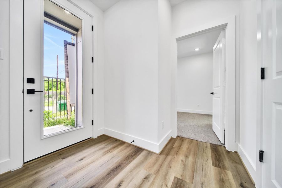 Bright entryway with modern hardwood flooring and a large glass door leading outside. The hallway opens to a carpeted room, giving a cozy feel.