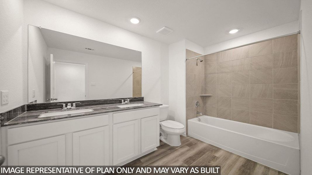 Bathroom featuring a dual vanity with a dark countertop and two undermount sinks, white cabinetry, a large wall-mounted mirror, a toilet, and a combination tub and shower with light-toned tiled walls