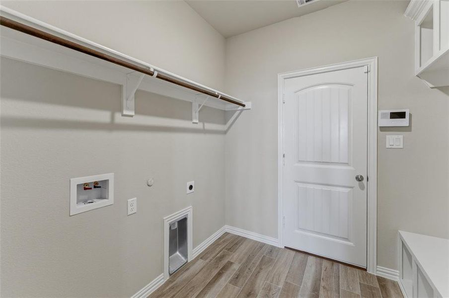 Dedicated laundry area featuring wood-finish flooring, light neutral wall tones, a built-in shelf with hanging rod, and utilities hookups
