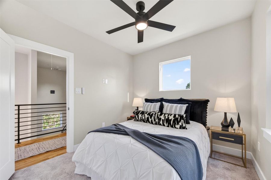 The secondary bedroom opens onto the third-floor landing featuring the home's signature black metal railing system. Plush carpet, neutral walls, and a ceiling fan make this a warm and restful room.