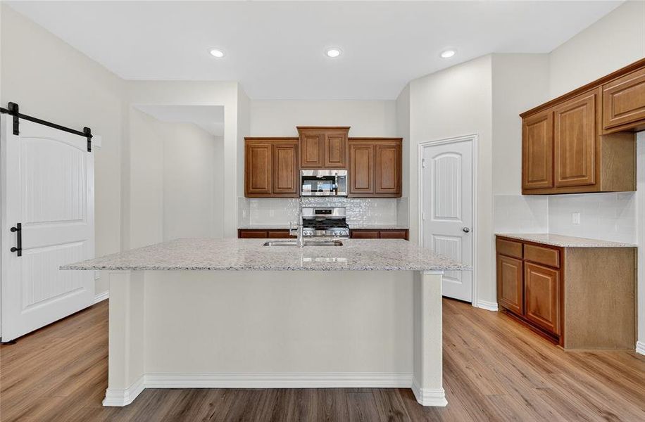 Kitchen with brown cabinetry, a barn door, tasteful backsplash, light stone counters, and recessed lighting