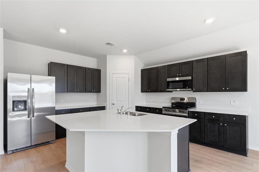 Kitchen featuring a central island with an undermount double-basin sink, stainless steel appliances, dark wood-finish cabinetry, light countertops, and wood-finish flooring Kitchen featuring a central island with an undermount double-basin sink, stainless steel appliances, dark wood-finish cabinetry, light countertops, and wood-finish flooring