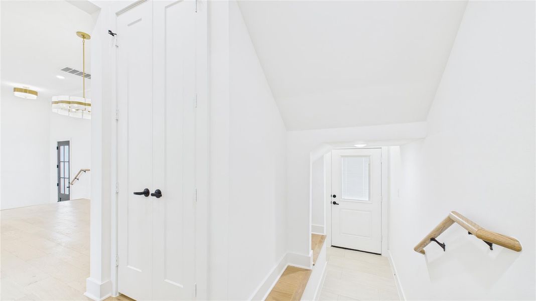 Bright and modern hallway with white walls and light wood floors. Features a staircase with a wooden handrail, a double door closet, and stylish light fixtures.