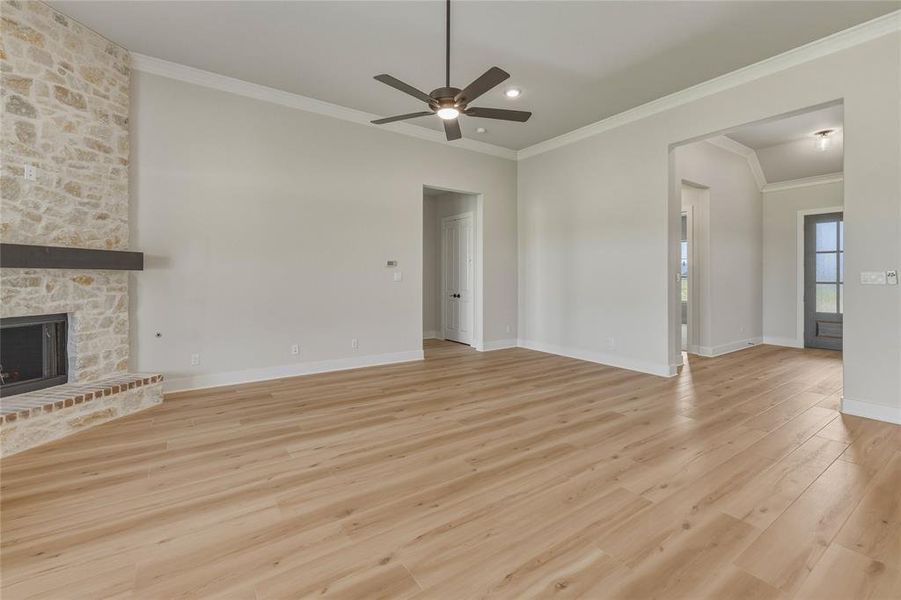Open-concept living area featuring light wood-finish flooring, a stone-clad fireplace with a wood mantel, crown molding, and a ceiling fan Open-concept living area featuring light wood-finish flooring, a stone-clad fireplace with a wood mantel, crown molding, and a ceiling fan