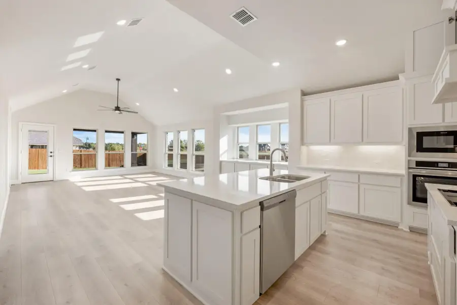 Open kitchen with white cabinets, large island, stainless appliances, and vaulted ceiling open to living area with light wood floors