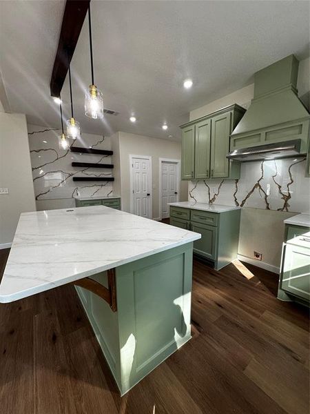 Kitchen featuring a central island with a waterfall countertop, sage green cabinetry, a matching range hood, wood-finish flooring, and a patterned full-height backsplash