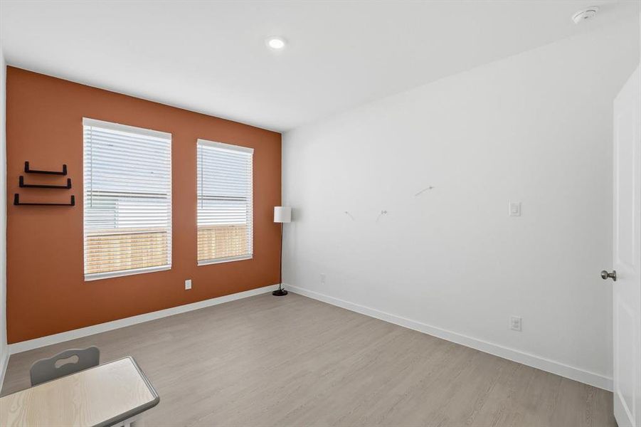 Bedroom featuring a warm accent wall, two window openings with blinds, and light wood-finish flooring Bedroom featuring a warm accent wall, two window openings with blinds, and light wood-finish flooring