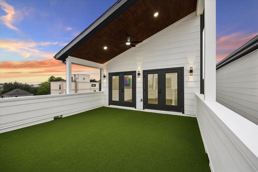 This photo depicts a modern rooftop terrace featuring artificial grass, a wooden ceiling with recessed lighting and a fan, and double doors leading inside. The space offers a stylish outdoor area with a sunset view. This photo depicts a modern rooftop terrace featuring artificial grass, a wooden ceiling with recessed lighting and a fan, and double doors leading inside. The space offers a stylish outdoor area with a sunset view.