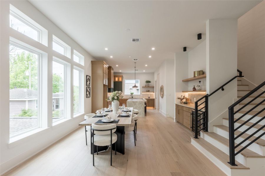 A wall of windows does the heavy lifting here. Three tall expanses of glass flood the dining footprint with borrowed treetop light, while the kitchen sits on-axis at the far end; a long gallery of oak, stone, and air.