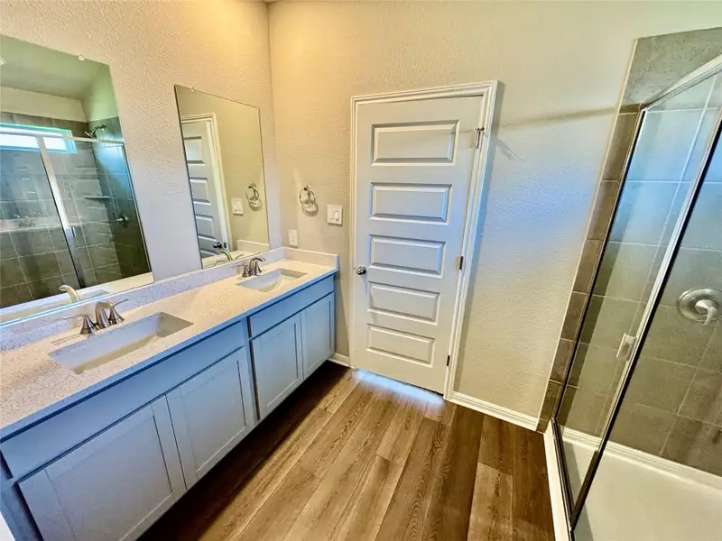 Bathroom featuring a textured wall, wood finished floors, a shower stall, and double vanity Bathroom featuring a textured wall, wood finished floors, a shower stall, and double vanity