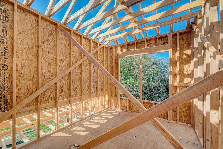 This photo shows a room under construction with wooden framing and a pitched roof. There's a large window opening with a view of trees, allowing natural light to fill the space.