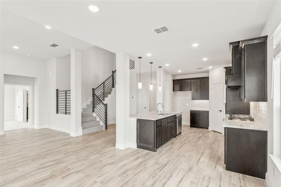 Open-concept kitchen with dark cabinets, center island, and light wood floors, open to living area with staircase visible Open-concept kitchen with dark cabinets, center island, and light wood floors, open to living area with staircase visible