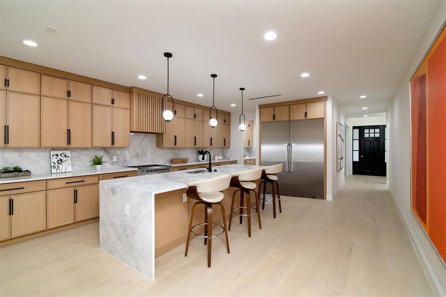 Kitchen with light wood-type flooring, light stone counters, stainless steel appliances, decorative backsplash, and a kitchen island with sink