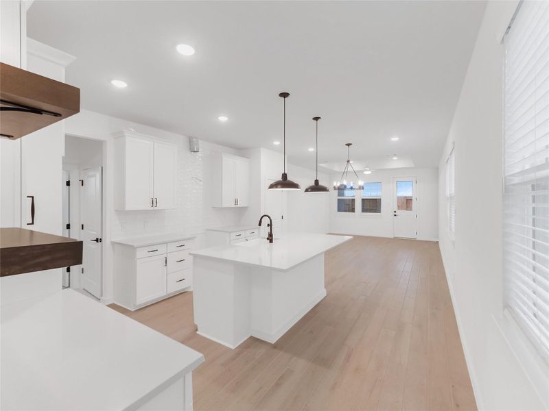 This spacious, modern kitchen features sleek white cabinetry, a large central island with a sink, and stylish pendant lighting. The open layout flows into a bright dining area with wood vinyl floors and ample natural light from multiple windows.