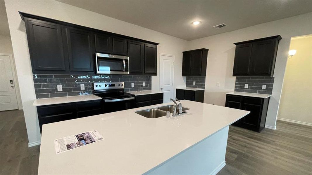 Kitchen featuring a large central island with an undermount double sink, dark cabinetry, light countertops, and a gray subway tile backsplash Kitchen featuring a large central island with an undermount double sink, dark cabinetry, light countertops, and a gray subway tile backsplash
