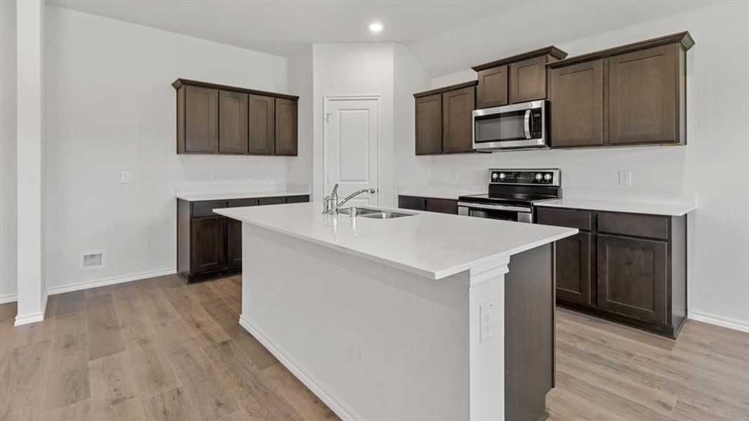 Kitchen featuring an island with a double basin stainless steel sink, light-toned countertops, wood-finish flooring, dark wood cabinetry, and stainless steel appliances