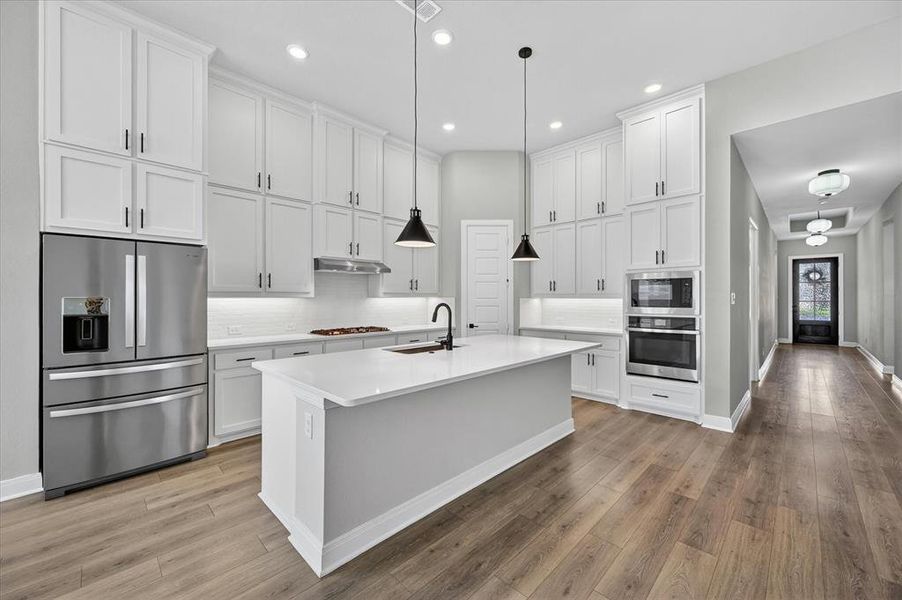 Kitchen featuring white shaker-style cabinetry, stainless steel appliances, a center island with a sink, and wood-finish flooring Kitchen featuring white shaker-style cabinetry, stainless steel appliances, a center island with a sink, and wood-finish flooring