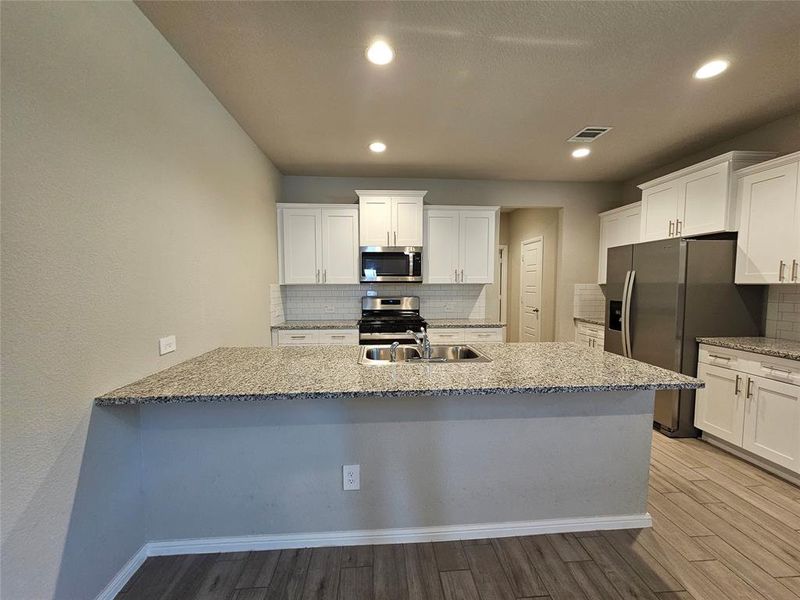 Kitchen with white cabinetry, open floor plan, stainless steel appliances, recess lights, granite counters, backsplash and wood looking tiled floors.
