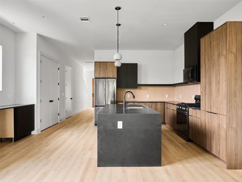 Kitchen featuring modern cabinets, an island with sink, black appliances, pendant lighting, and light wood-style floors Kitchen featuring modern cabinets, an island with sink, black appliances, pendant lighting, and light wood-style floors
